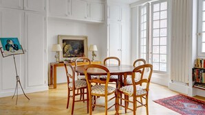 A dining room with a wooden table and chairs, a painting on the wall, and a bookshelf.