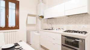 Une cuisine compacte avec des meubles blancs, un four encastré, une cuisinière à gaz et une fenêtre équipée de stores.