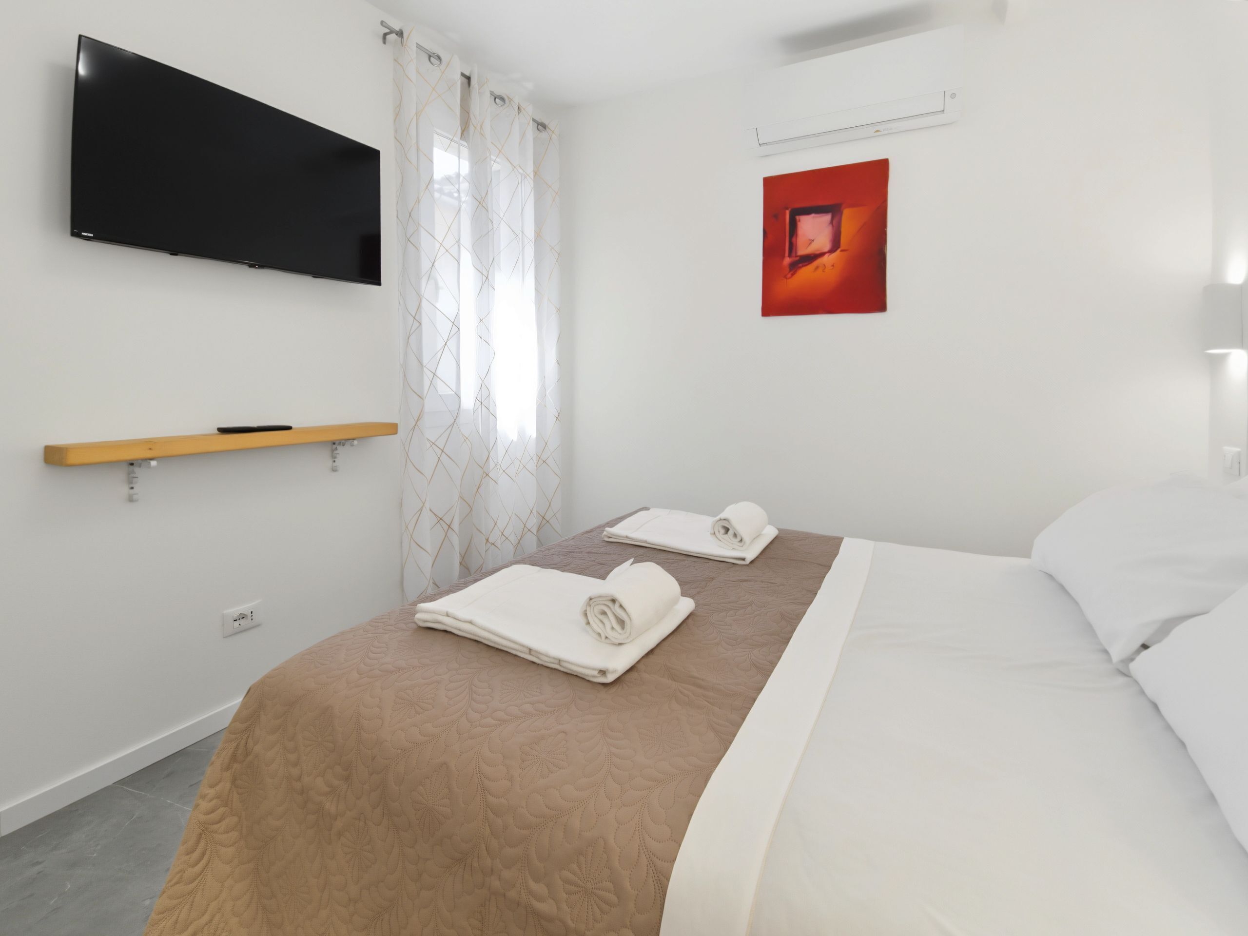 A hotel room with a bed, a flat-screen TV mounted on the wall, a red artwork, and a window with curtains.