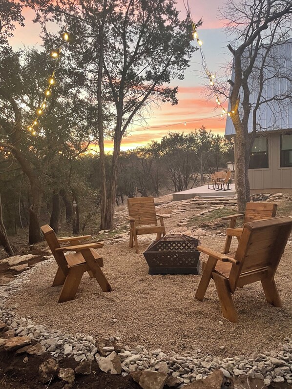 An outdoor seating area with wooden chairs and a fire pit, surrounded by trees and a sunset view. - Pet-friendly retreat in Granbury with loft, fire pit, and boat launch 2 mi away. (Granbury)