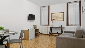 A living room with a sofa, a dining table with chairs, a TV mounted on the wall, and a window with curtains. - Rialto Dream (Venezia)