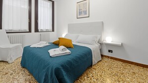 A hotel room with a bed, two windows with curtains, a radiator, a white chair, a bedside table with a lamp, and a framed picture on the wall. - Rialto Dream (Venezia)