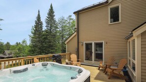 Una vasca idromassaggio su un pontile di legno, con sedie e un barbecue, circondata da alberi e una casa.