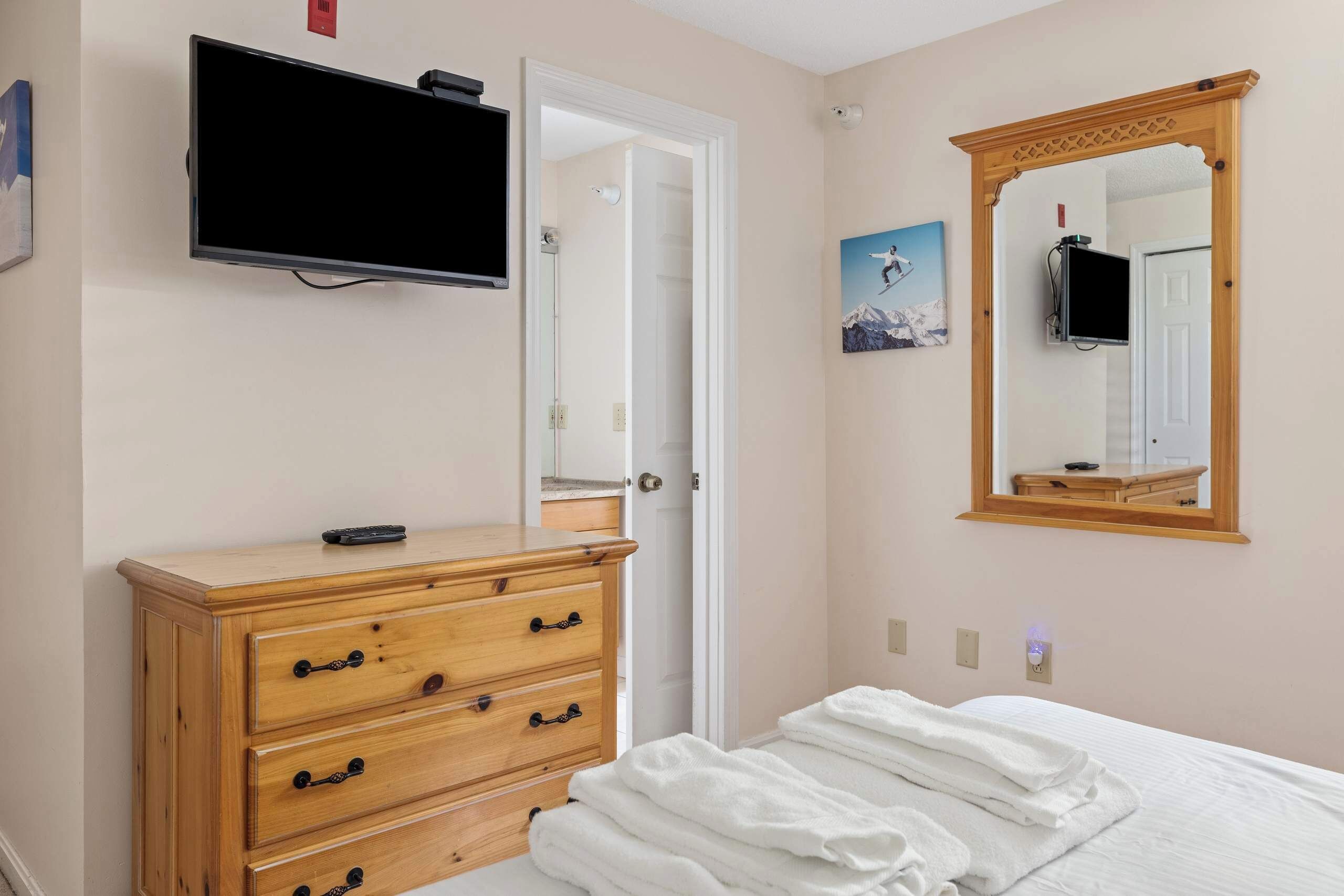 A bedroom with a bed, a wooden dresser, a television mounted on the wall, and a mirror.