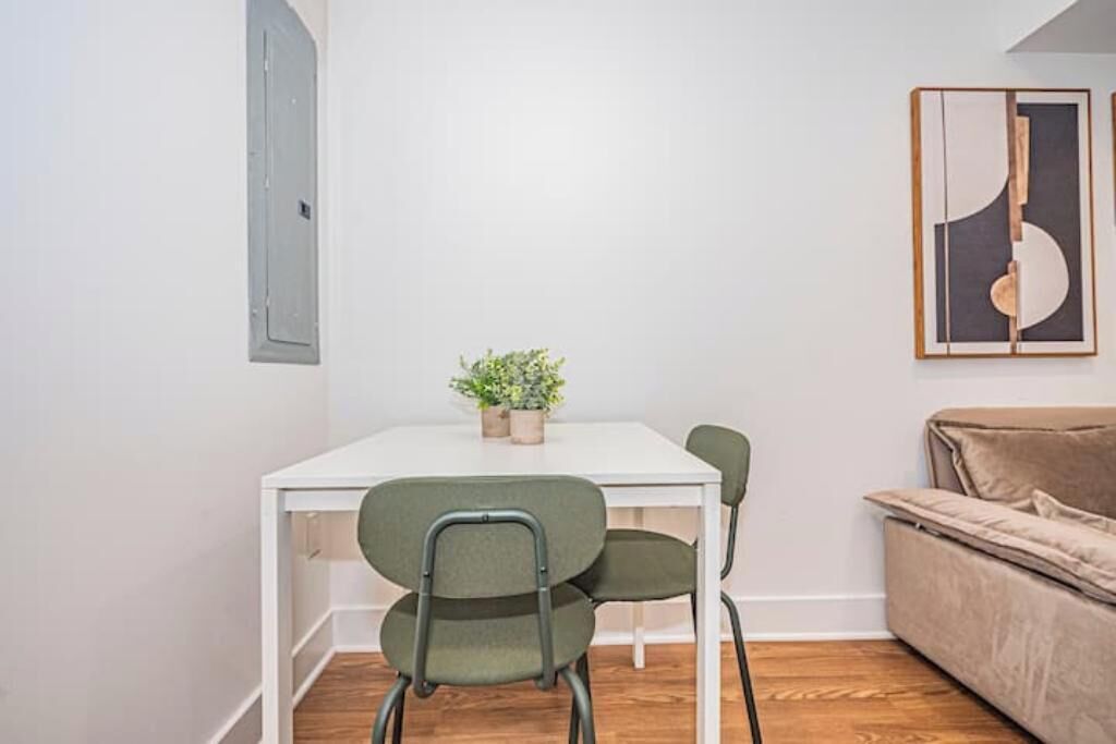 A small, modern room with a white table, two green chairs, a beige sofa, and a framed abstract art piece on the wall.