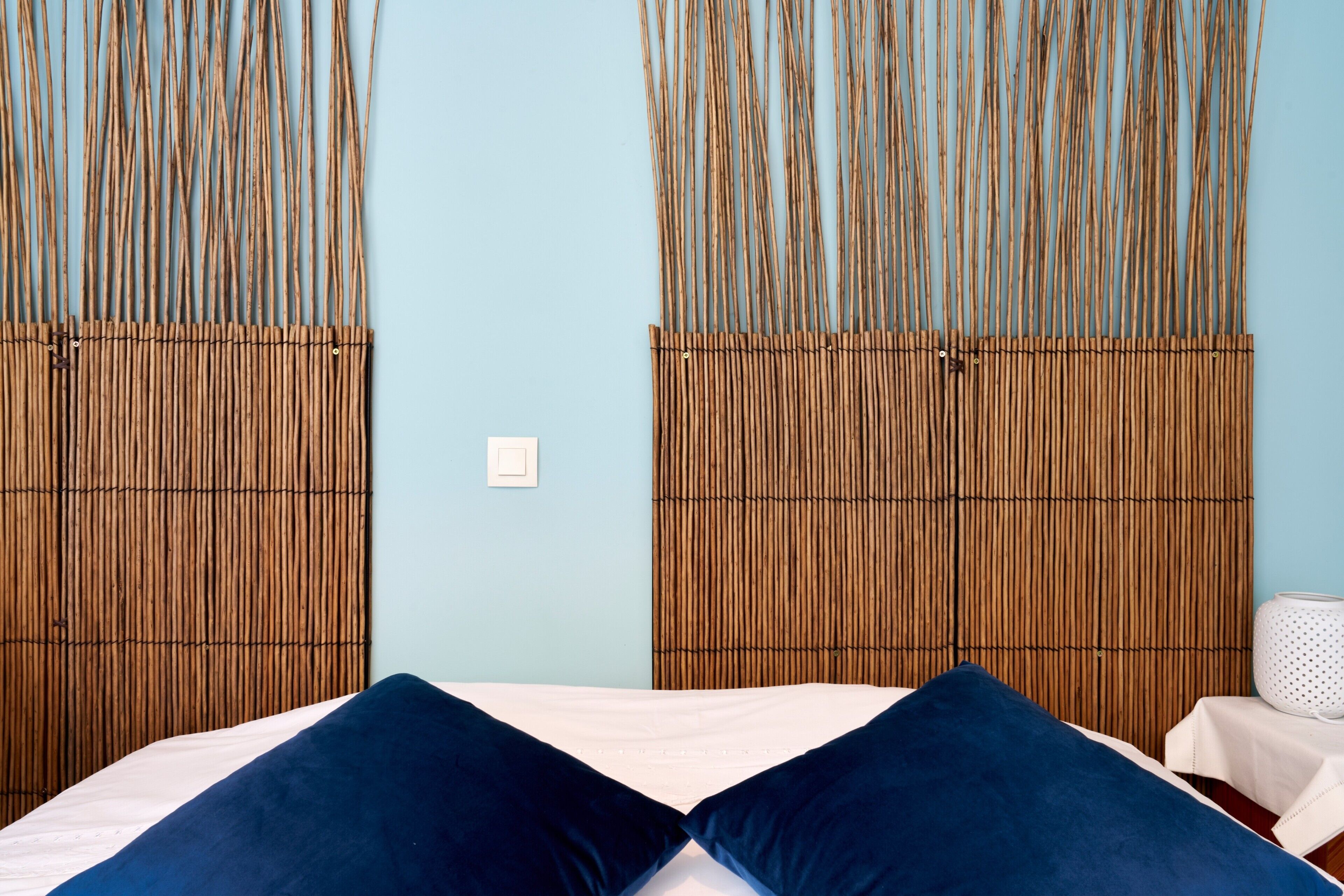 Une pièce comprenant un lit, deux panneaux de bambou verticaux, une table blanche recouverte d’une nappe blanche et une lampe blanche.