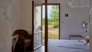 Quarto com cama, cadeira, mesa, ventilador e vista para o exterior.
