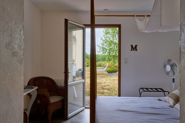 A bedroom with a bed, a chair, a table, a fan, and a view of the outdoors.