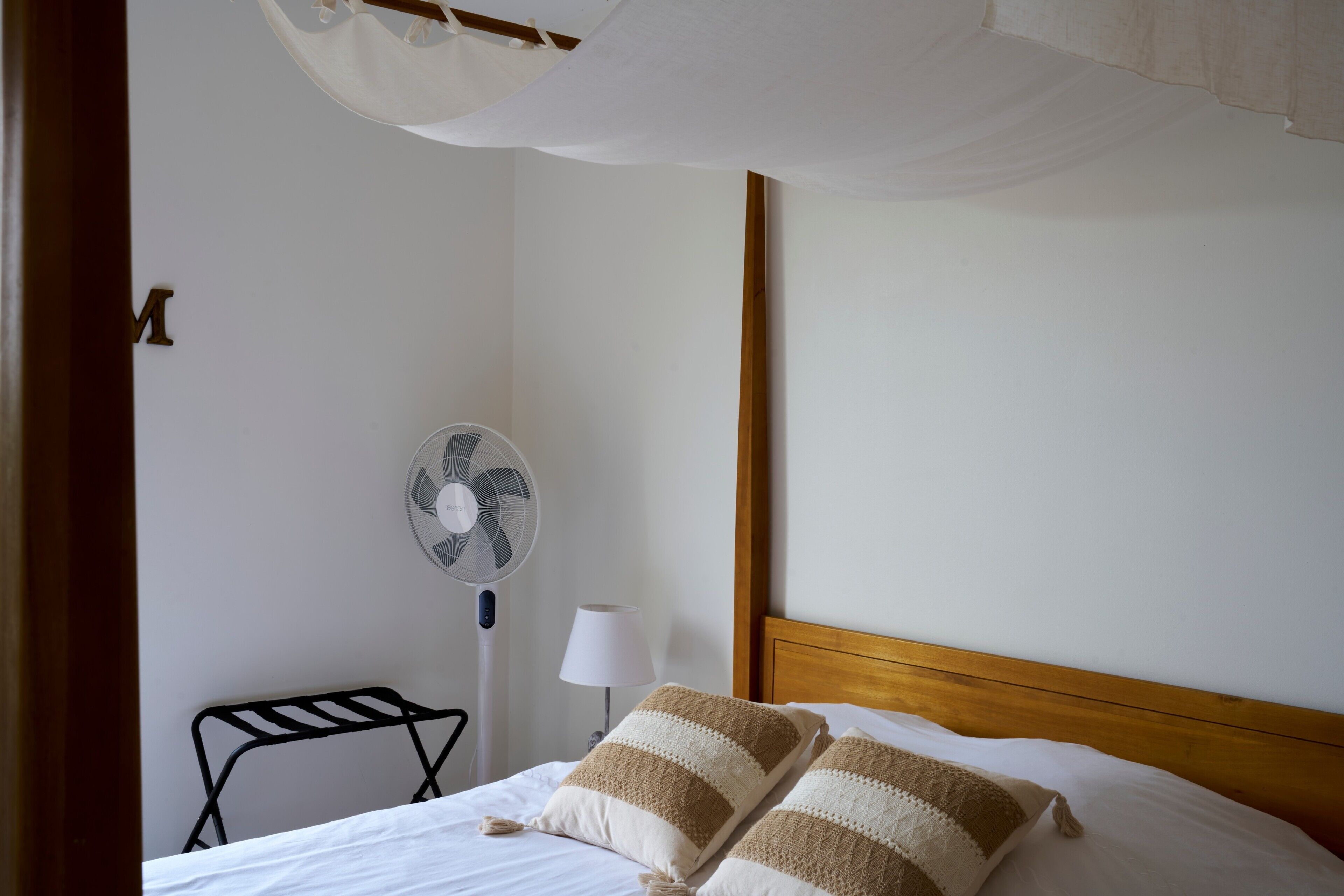 Una cama con almohadas a rayas, un ventilador de pie, una lámpara de noche y un cabecero de madera.