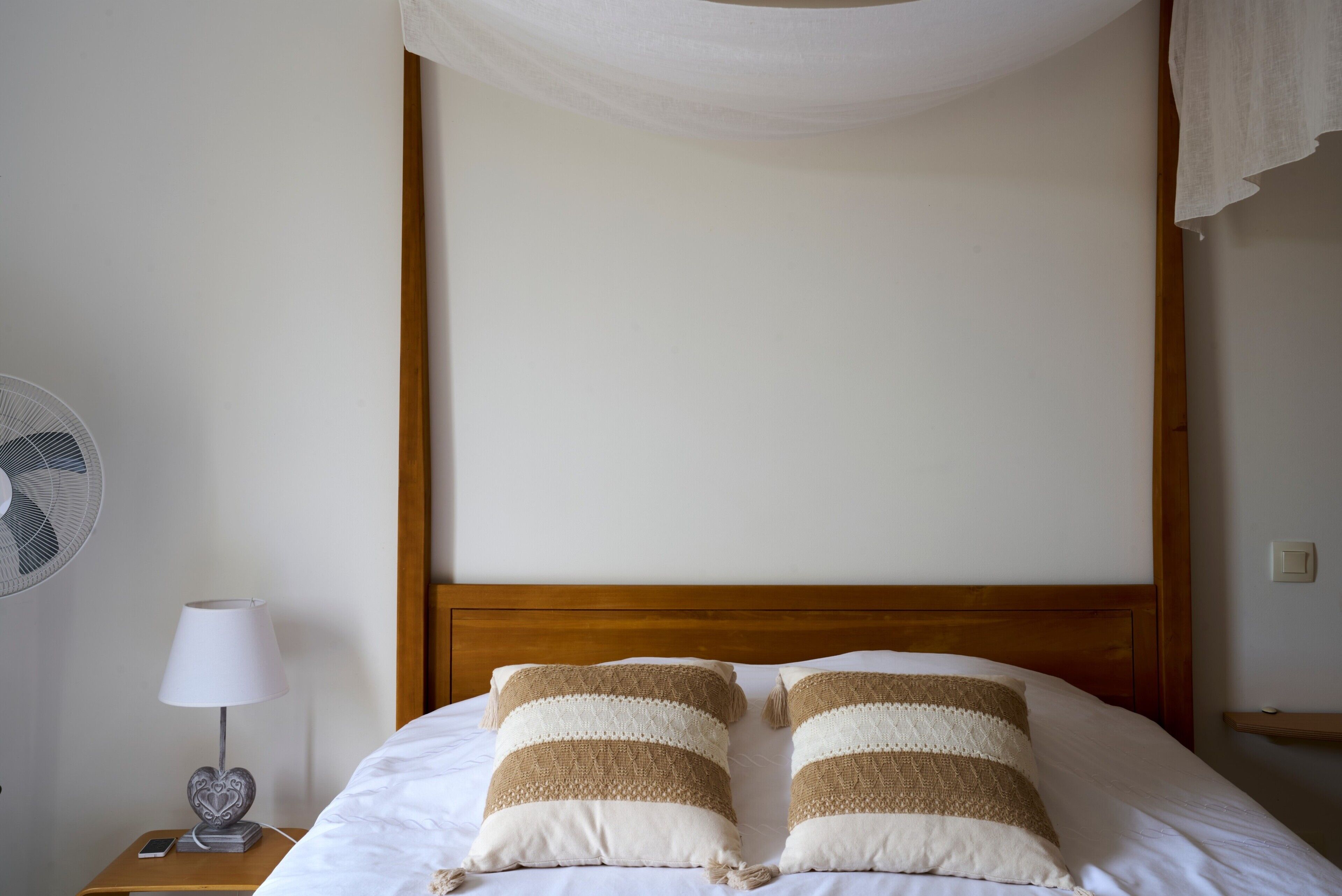 A bed with two striped pillows, a wooden headboard, a bedside table with a lamp, and a wall-mounted fan.