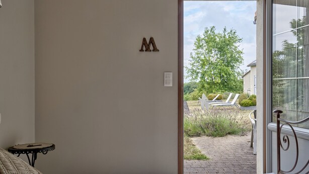 Une chambre à coucher avec un lit, une petite table, la lettre « M » peinte sur le mur, et une vue sur un jardin avec des chaises et une clôture blanche.
