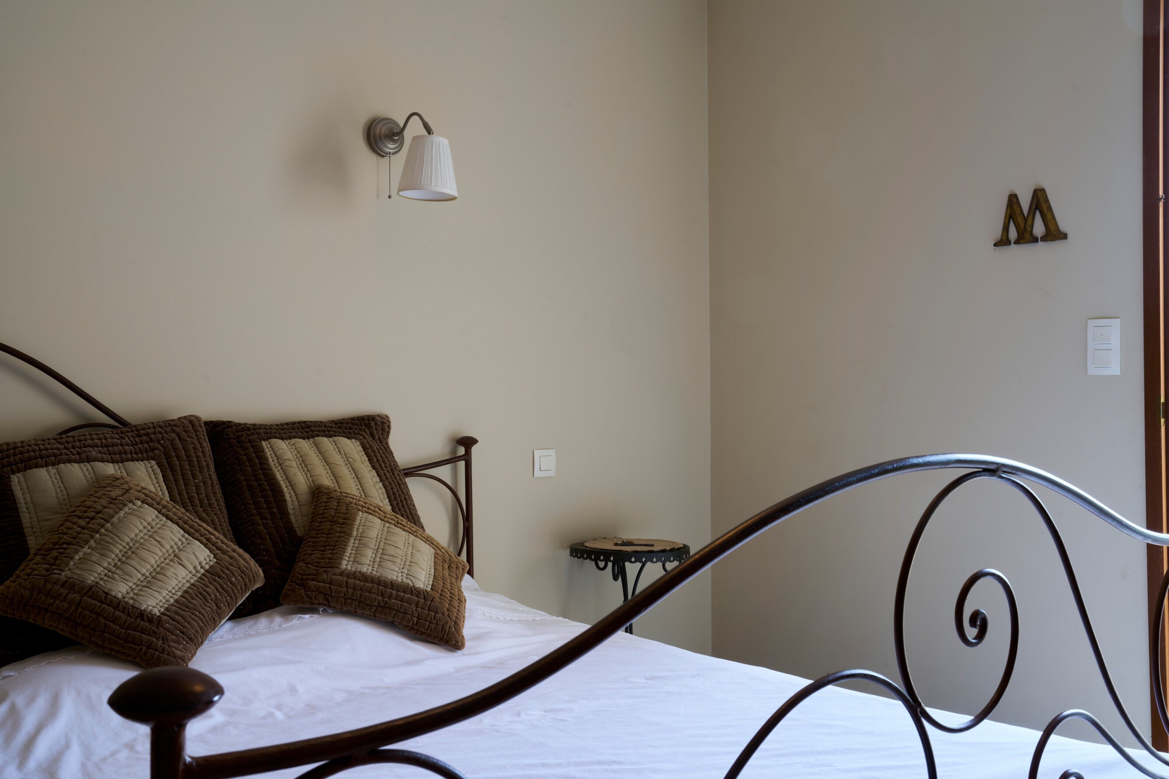 Un llit amb capçalera, una làmpada de paret, una taula petita i una decoració amb la lletra 'M' a la paret.