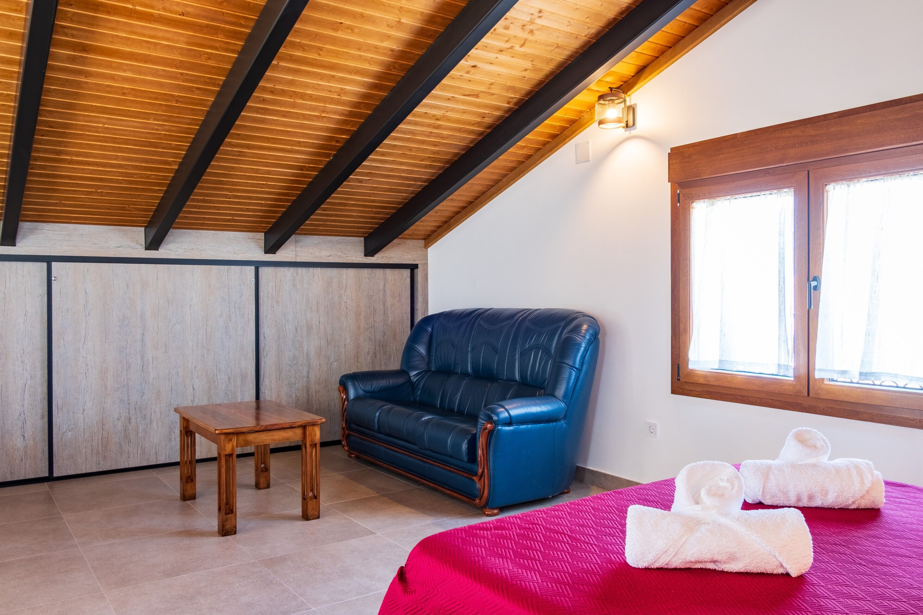 A room with a wooden ceiling, a blue leather sofa, a small wooden table, and a bed with a red bedspread.