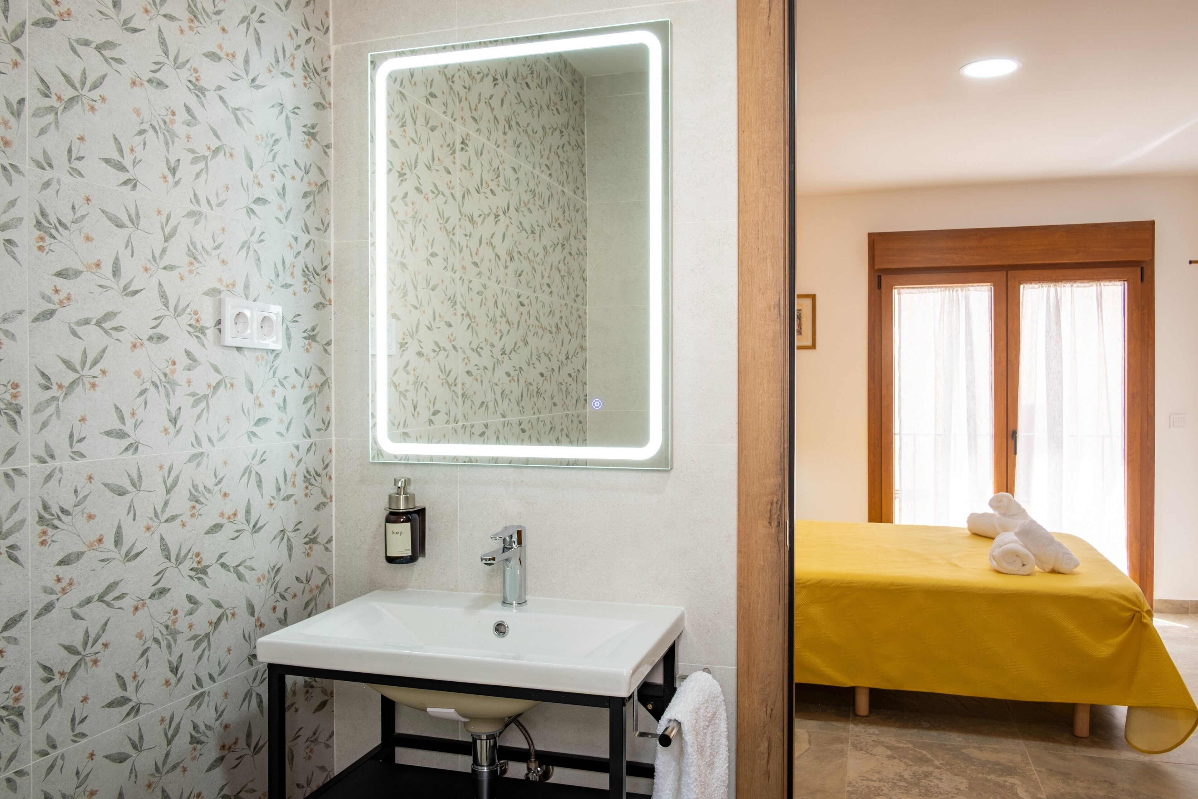 A bathroom with a floral wallpaper, a white sink, and a mirror with a light.