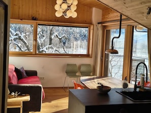 Une pièce chaleureuse avec un plafond en bois, une grande fenêtre donnant sur des arbres couverts de neige, un canapé, une table à manger et un coin cuisine.