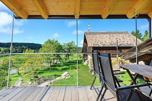 Une terrasse en bois offrant une vue sur un paysage rural, avec notamment une maison dotée d’un toit en tuiles et un mât pour drapeau.