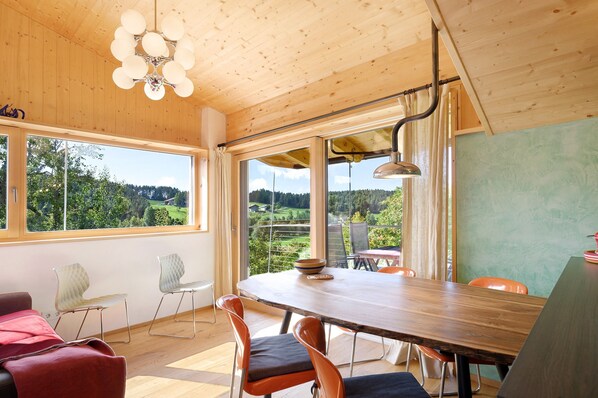 Un espace repas avec une table en bois, des chaises et une vue sur un paysage verdoyant, visible à travers de grandes fenêtres.