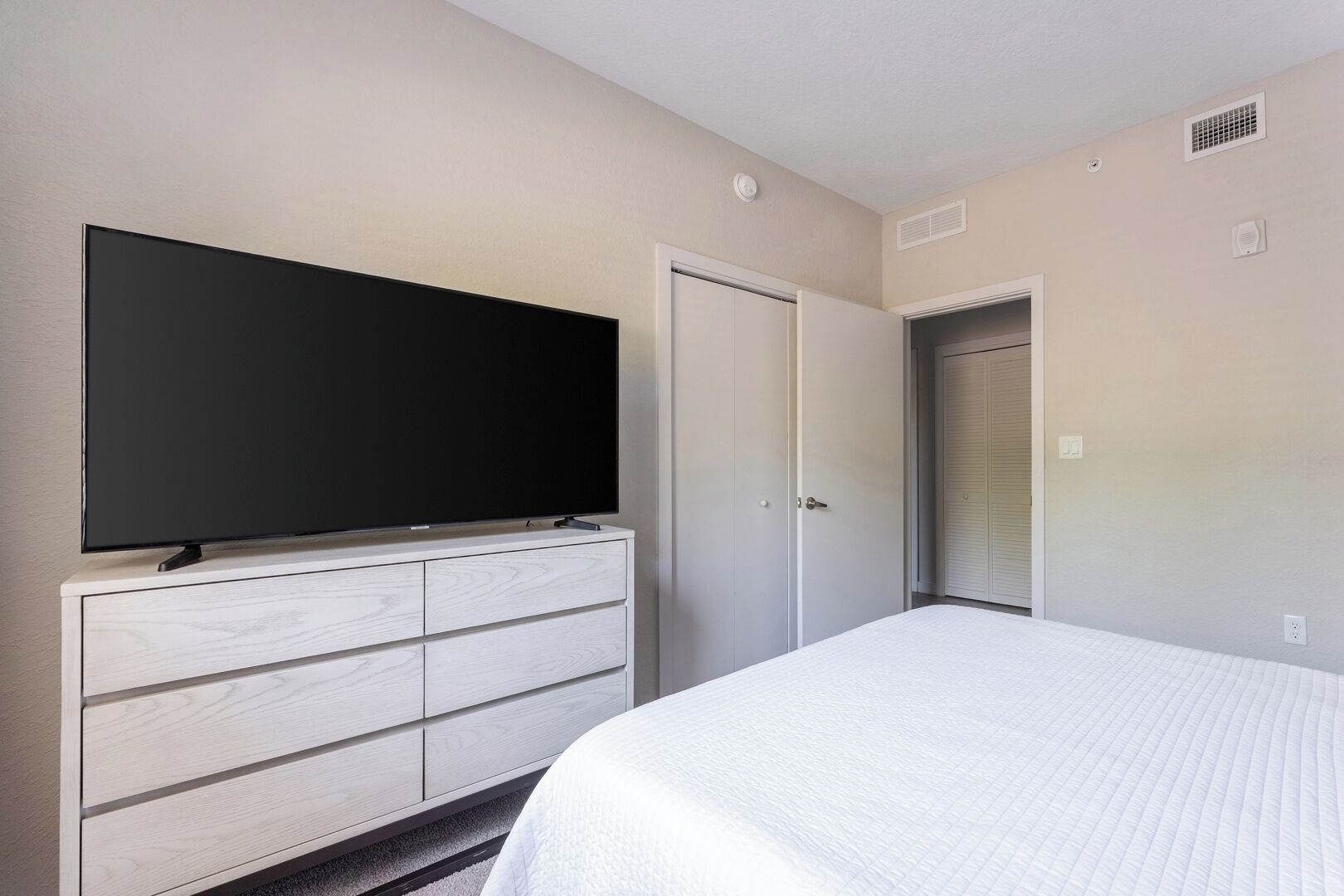 A hotel room with a large flat-screen TV mounted on a white dresser, a bed with white bedding, and a closed door.