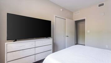 A hotel room with a large flat-screen TV mounted on a white dresser, a bed with white bedding, and a closed door.