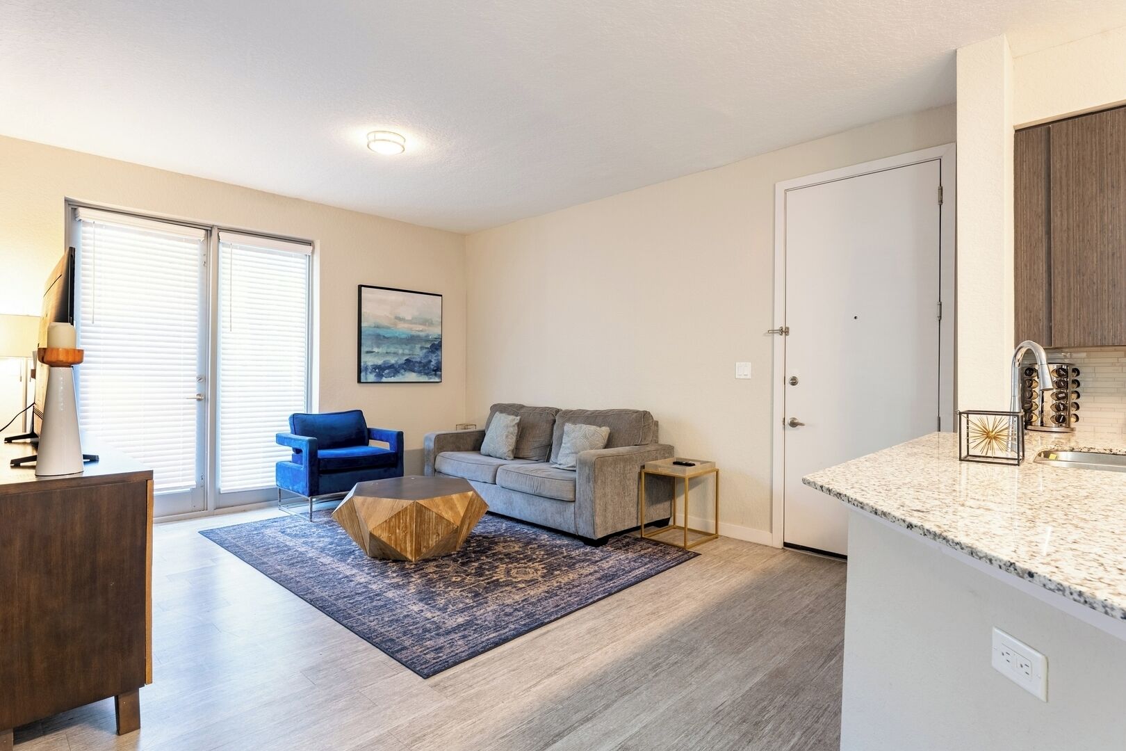 A modern living room with a grey sofa, a blue armchair, and a coffee table. There is a kitchen area with a granite countertop and a sink.