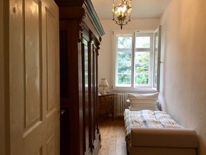 Um corredor estreito com porta de madeira, radiador, pequena mesa com abajur e cama com colcha listrada.