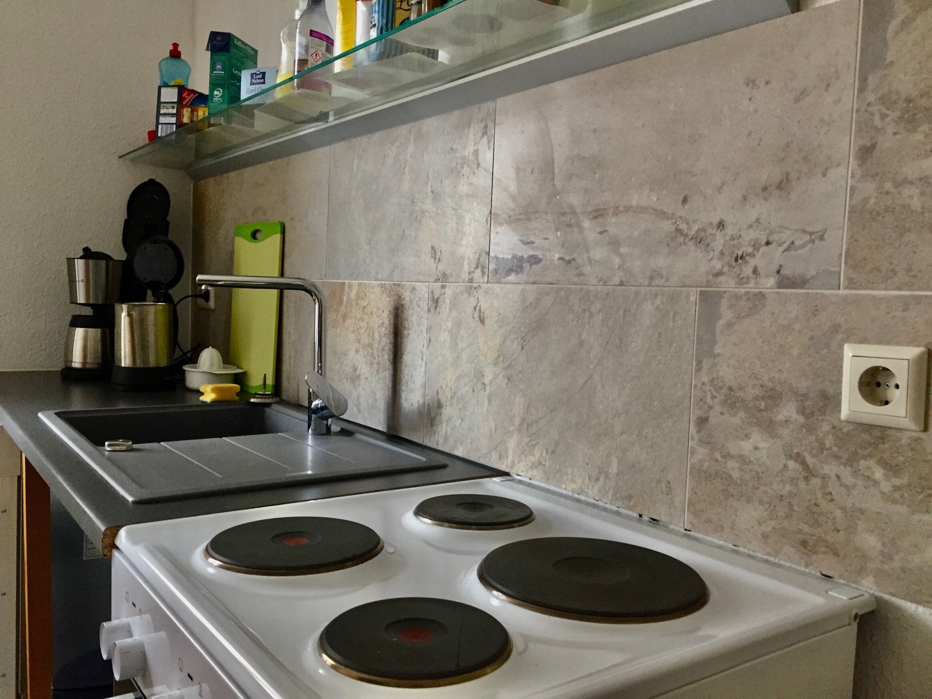 A kitchen with a stove, sink, and a shelf with various items.