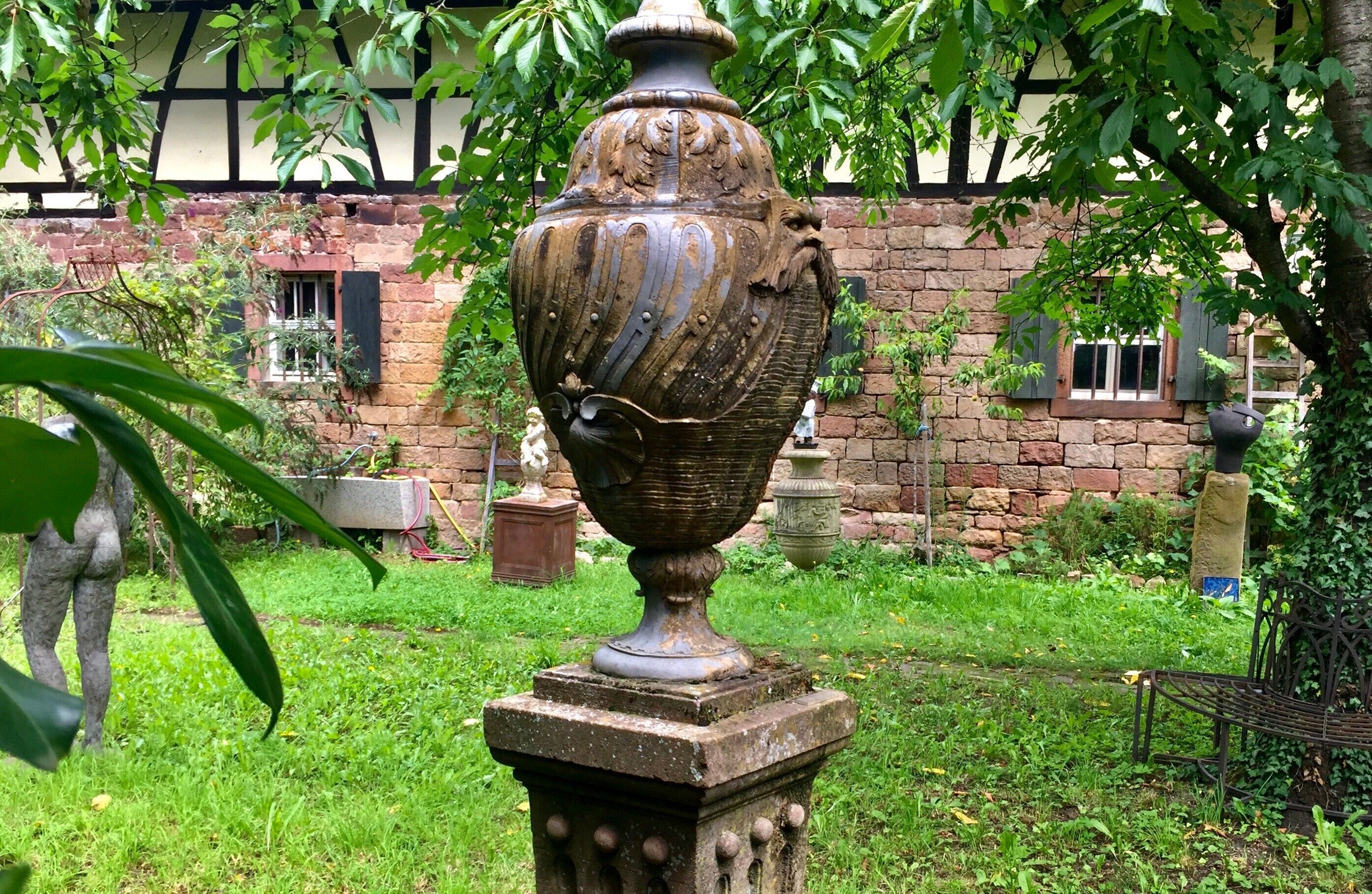 Een tuin met een grote urn op een voetstuk, omringd door groen en een gebouw met ramen.