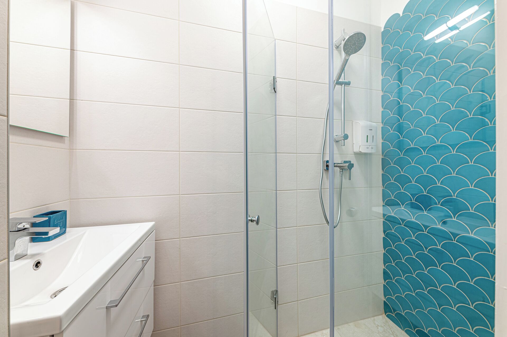 A bathroom with white tiles, a glass shower enclosure, and a sink with a blue soap dispenser.