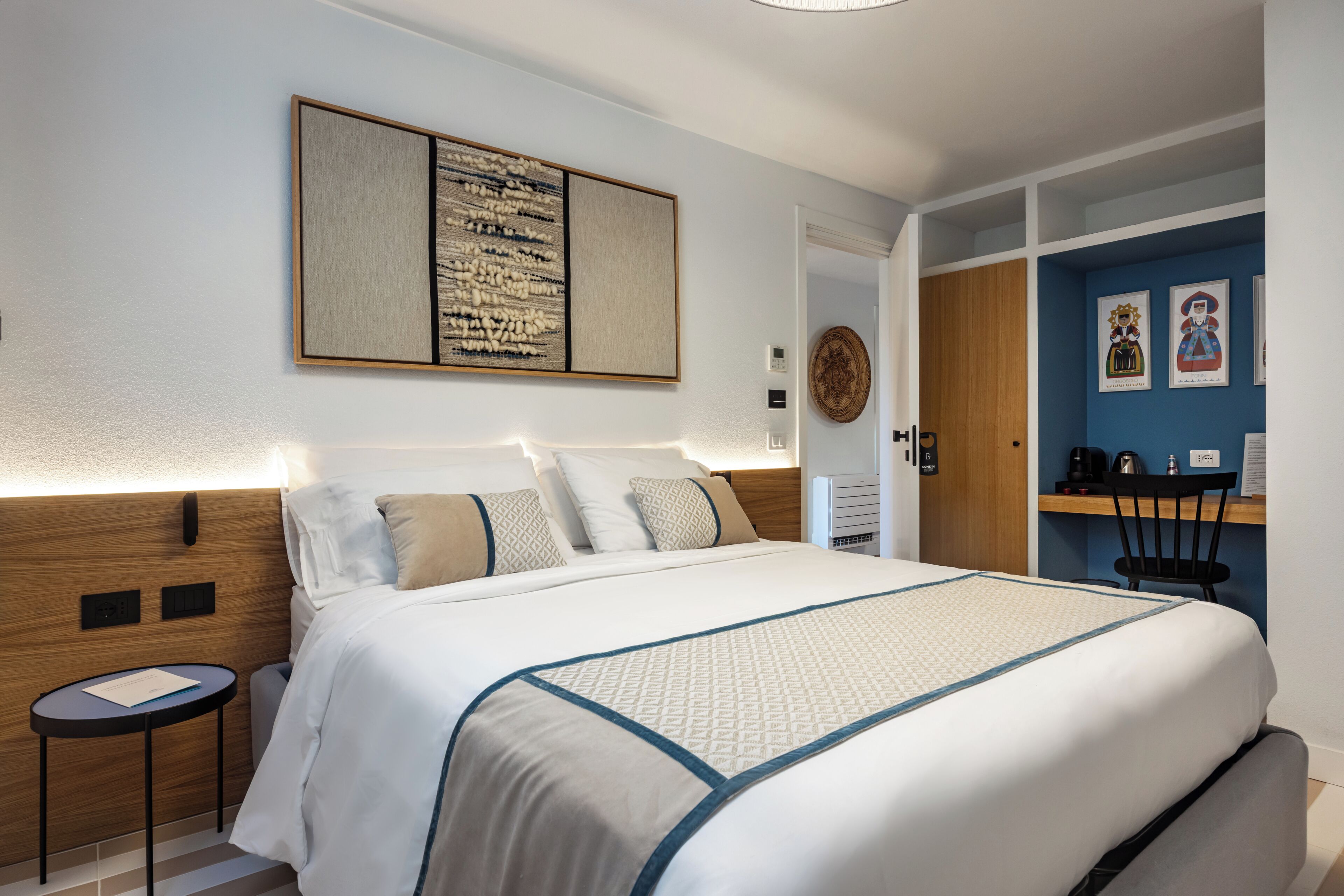 A hotel room with a large bed, a small side table, a desk with a chair, and a wall-mounted artwork.