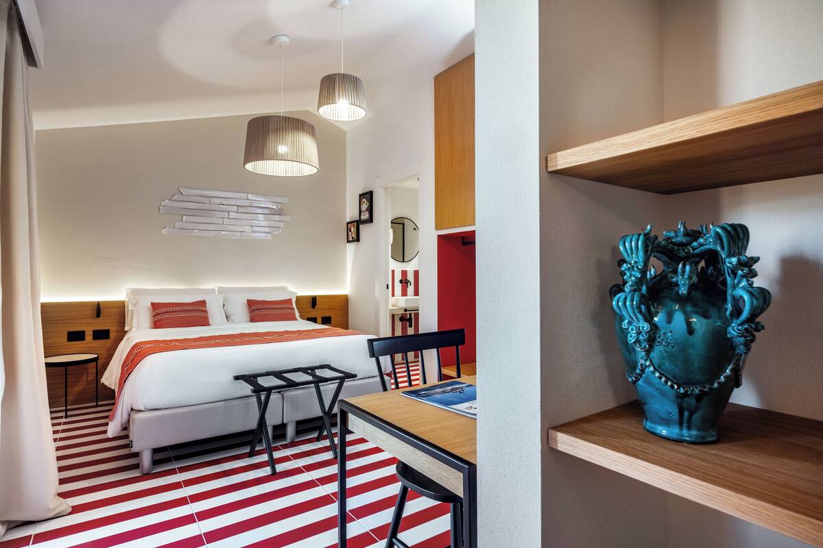 Una habitación de hotel con cama, mesita de noche, silla, escritorio y un jarrón decorativo.