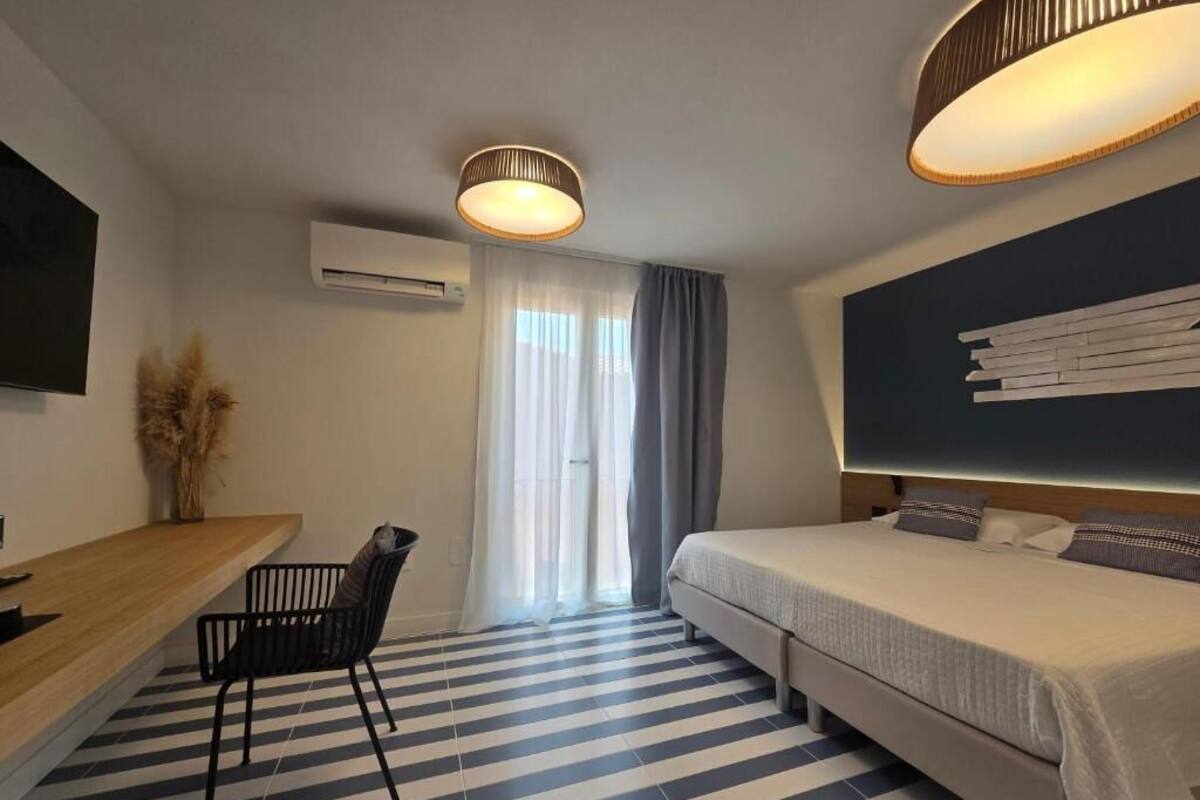 Una habitación de hotel con cama, escritorio con silla, ventana grande con cortinas, unidad de aire acondicionado y televisor montado en la pared.