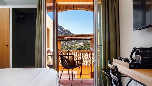 A hotel room with a bed, a desk with a chair, a TV, and a balcony with a view of the mountains. - Valesmeralda Boutique Rooms  (Arzachena)