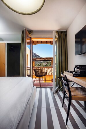 A hotel room with a bed, a desk with a chair, a TV, and a balcony with a view of the mountains. - Valesmeralda Boutique Rooms  (Arzachena)