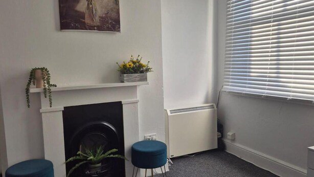 Een kamer met een witte open haard, een radiator, een raam met jaloezieën en twee blauwe krukken.