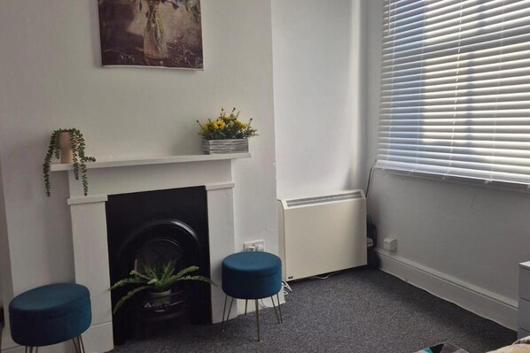 Een kamer met een witte open haard, een radiator, een raam met jaloezieën en twee blauwe krukken.