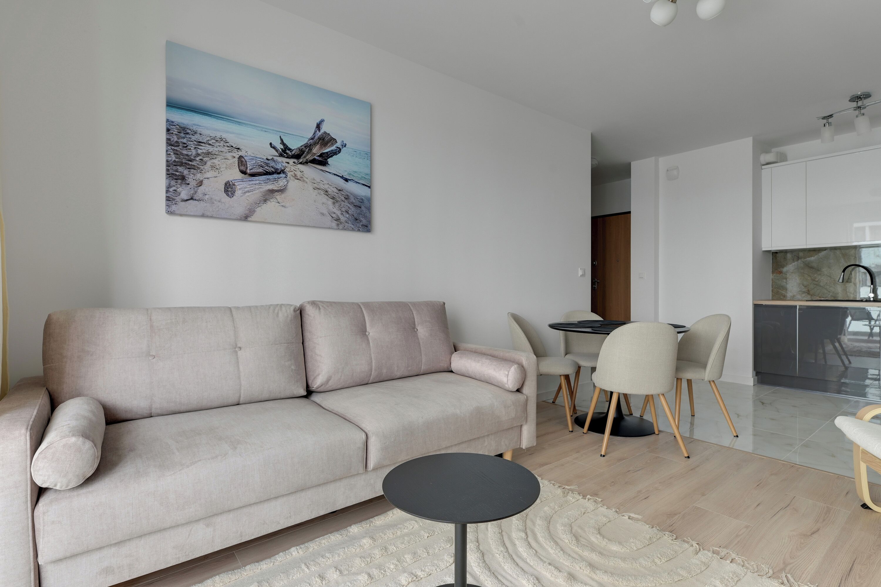 Ein Wohnzimmer mit einem beigefarbenen Sofa, einem runden schwarzen Couchtisch und einem Bild mit einer Strandansicht.