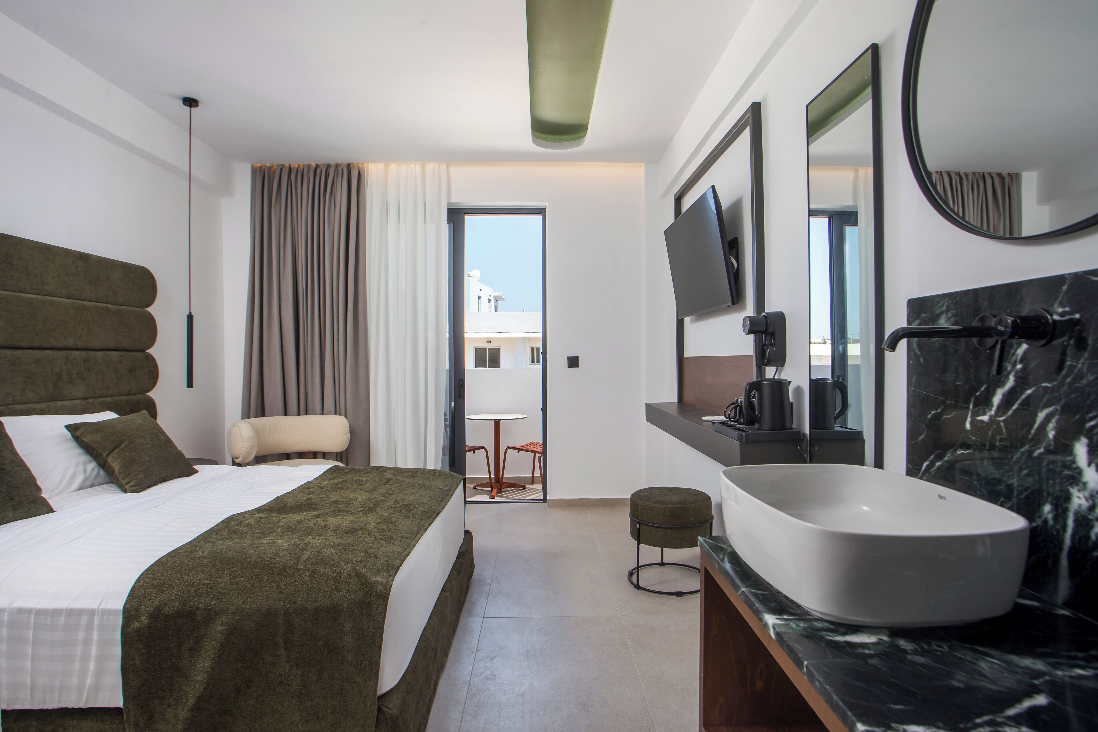 A modern hotel room with a large bed, a sitting area, a bathroom with a large sink, and a TV mounted on the wall.