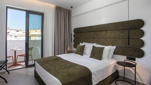 A modern hotel room with a large bed, a round side table, a chair, and a view of the city through a sliding glass door. - Ermou 44 City Hotel (Faliraki)