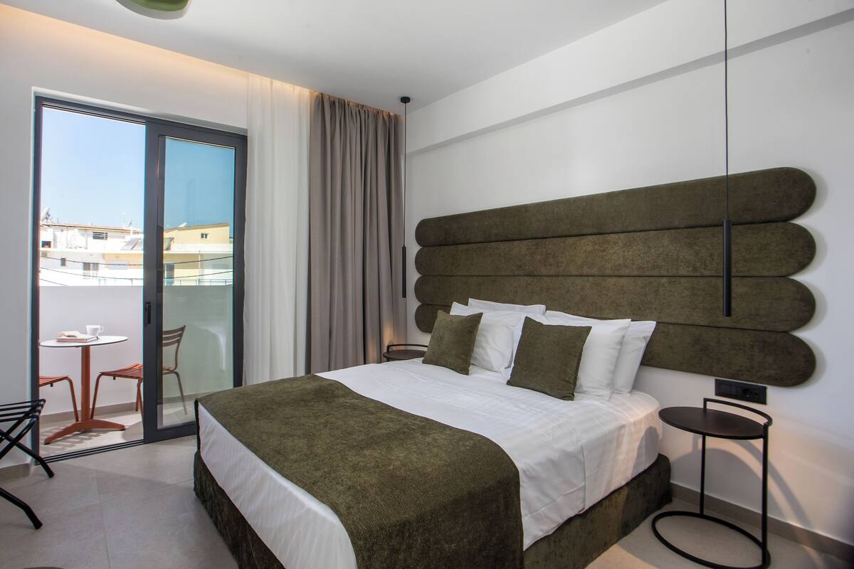 Moderni hotellihuone, jossa on suuri sänky, pyöreä sivupöytä, tuoli ja liukuovellinen lasiterassi, josta avautuu näkymä kaupunkiin.