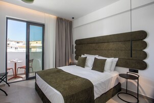 A modern hotel room with a large bed, a round side table, a chair, and a view of the city through a sliding glass door.