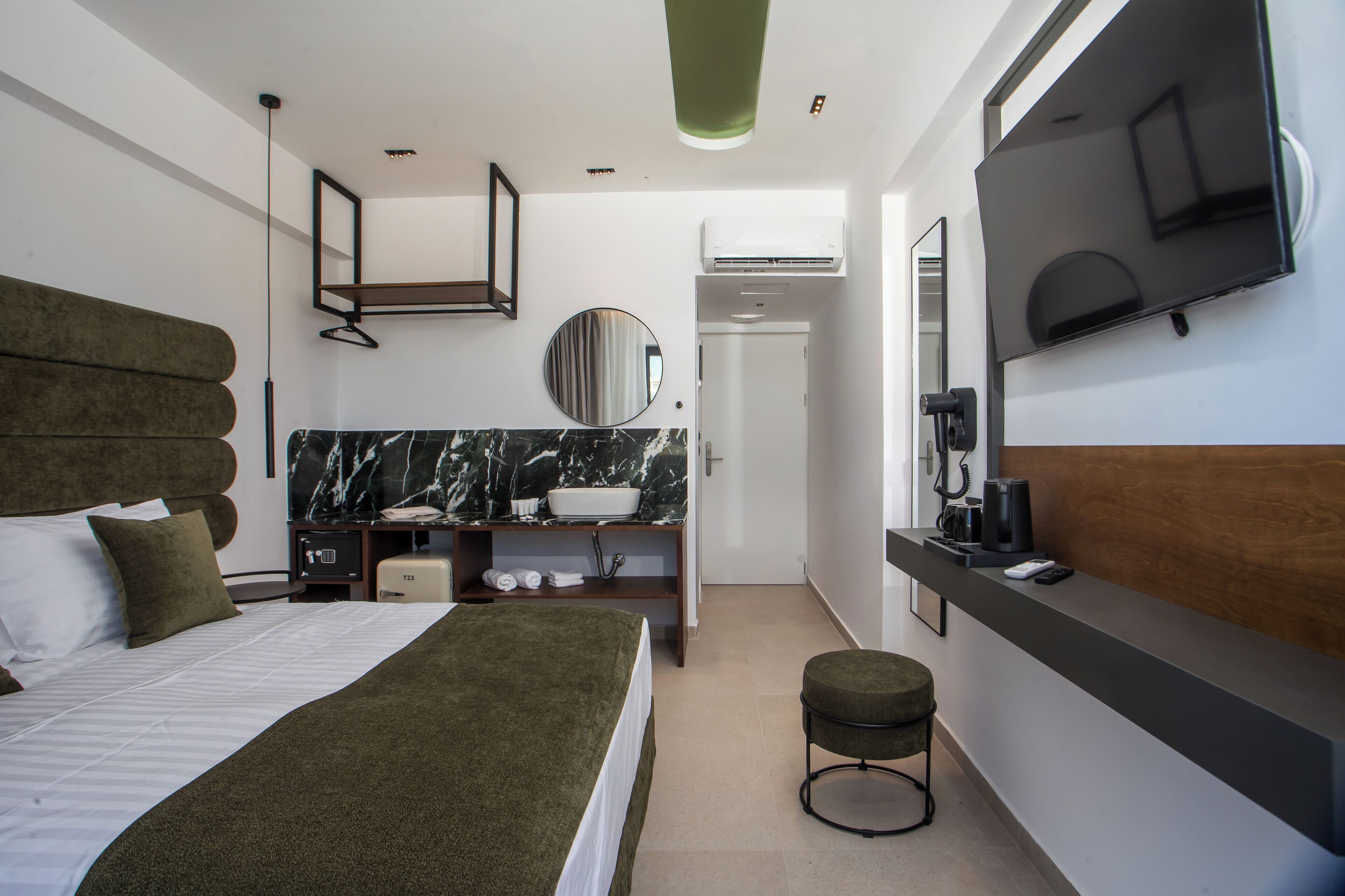 Quarto de hotel moderno com uma cama grande, uma penteadeira com espelho, uma televisão montada na parede e um banquinho pequeno.