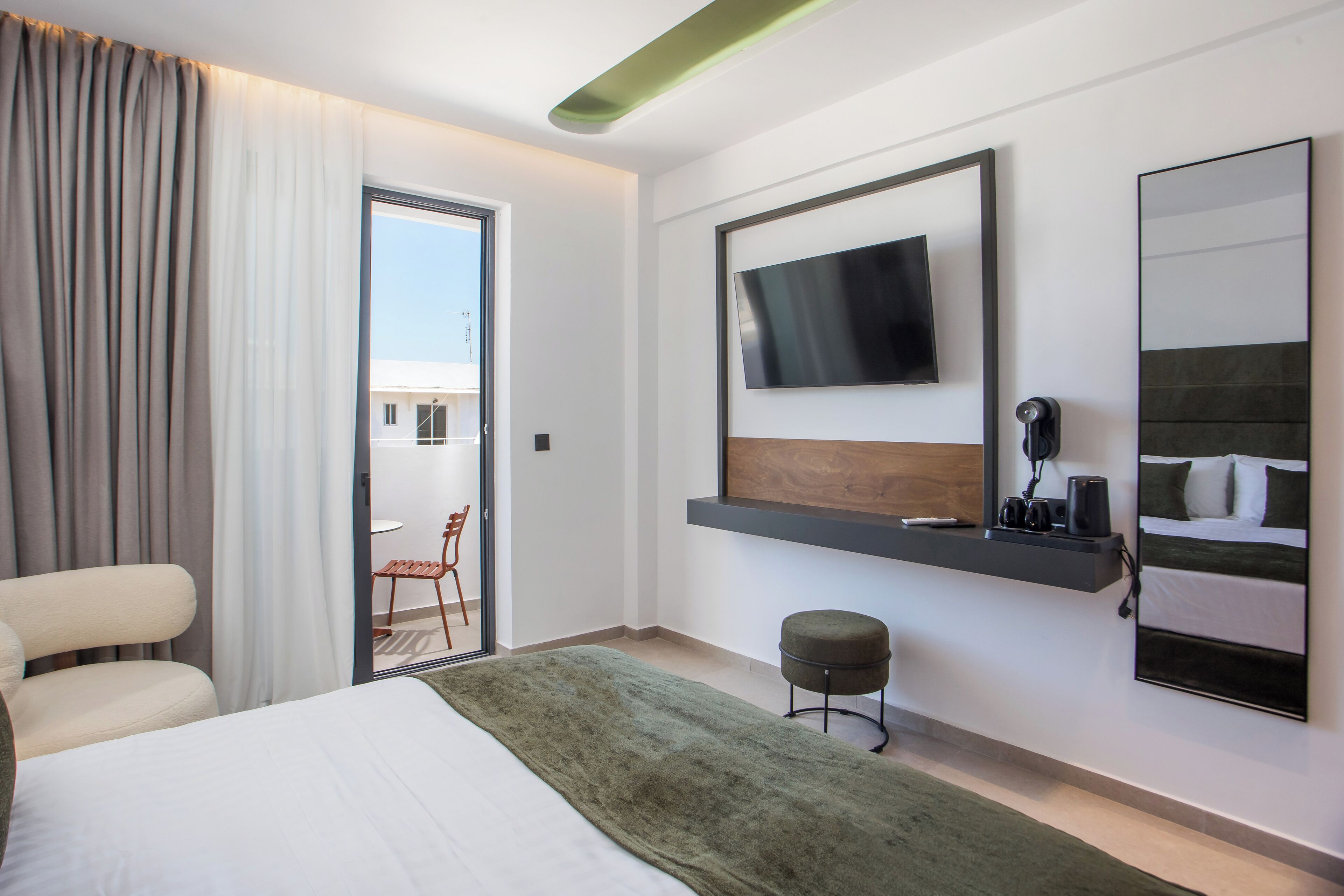 A modern hotel room with a bed, a seating area with a chair, a small table, a TV mounted on the wall, and a large window with curtains.