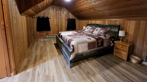 Une chambre à coucher aux murs lambrés, avec un plafond en pente, un lit recouvert d’une couvre-lit à motifs, une table de chevet avec une lampe et un plancher en bois.