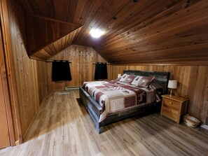Une chambre à coucher aux murs lambrés, avec un plafond en pente, un lit recouvert d’une couvre-lit à motifs, une table de chevet avec une lampe et un plancher en bois.