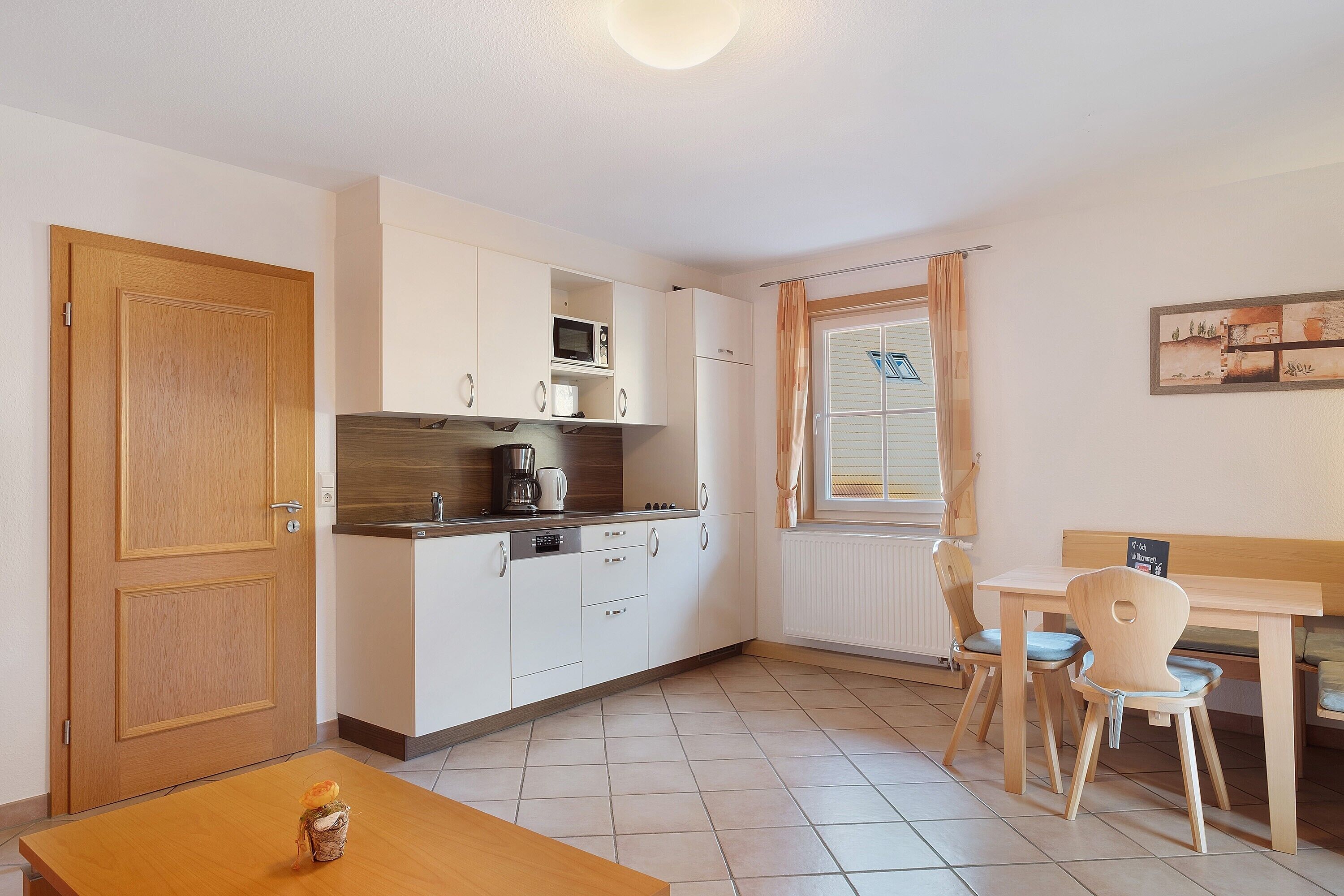 Een kleine keuken met witte kastjes, een magnetron en een koelkast. Er is een houten tafel met een stoel en een raam met gordijnen.