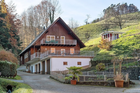 A two-story wooden house with a balcony, a stone wall, and a small garden.