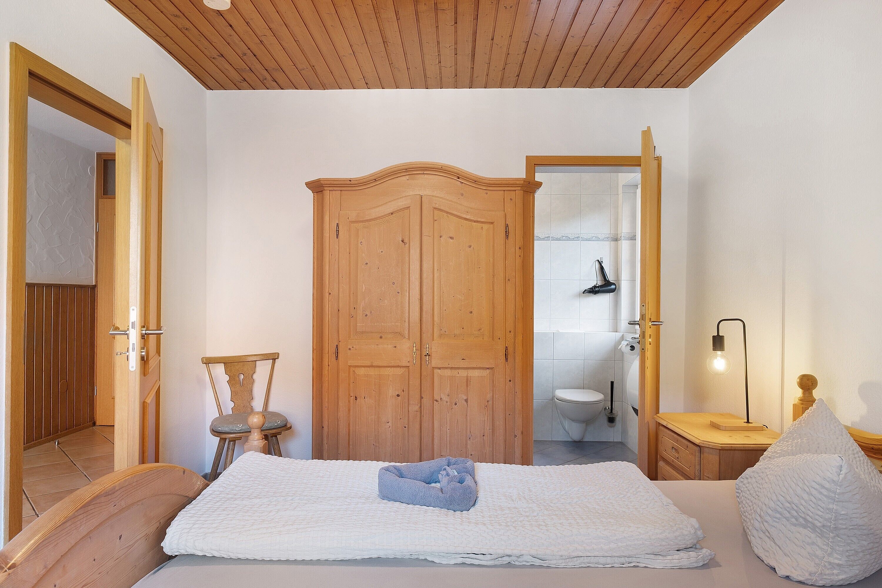 A bedroom with a wooden wardrobe, a bed with a white bedspread, a bedside table with a lamp, and a wooden chair.