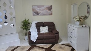 A living room with a brown sofa, a white throw blanket, a potted plant, a white dresser, a mirror, and a wall-mounted abstract painting.