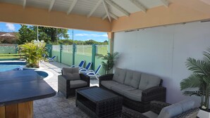 A covered patio area with wicker furniture, a pool, and a view of a green fence and trees.