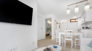 Una sala de estar moderna con un televisor de pantalla plana montado en la pared, una zona de comedor con una mesa y sillas blancas, y una cocina con armarios blancos y lámparas colgantes.